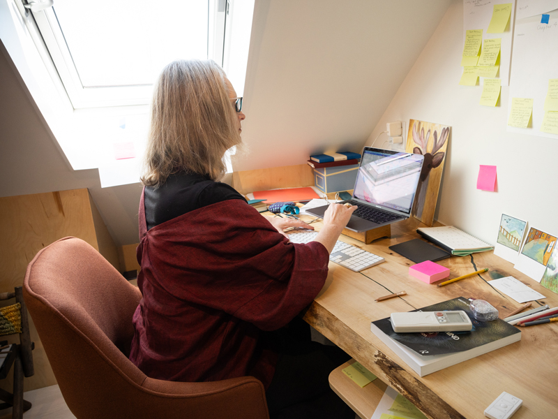 A photo of Kristin Andrews sitting at her desk and working on her laptop. The desk is situated in a corner by a window. There are sticky notes on the wall in front and on her desk, along with postcards, a keyboard and notebooks. Photo de Kristin Andrews assise à son bureau et travaillant à son ordinateur portable. Le bureau se trouve dans un coin, près d’une fenêtre. Il y a des notes adhésives sur le mur en face et sur son bureau, ainsi que des cartes postales, un clavier et des cahiers.